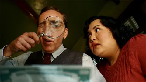 Aaron Epstein/Prime Character of Ben Reilly on the left wearing a brown waistcoat and maroon tie on a shirt, holding a magnifying glass, with his loyal secretary Janet, wearing a red top, looking at Reilly.