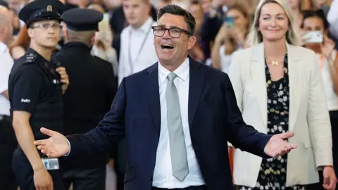 Dan Kitwood via Getty Images Andy Burnham wearing a navy blue suit, a grey tie and white shirt, holding his arms out.