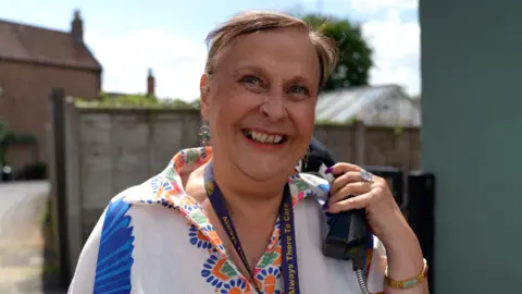 BBC / Crispin Rolfe Caroline Ali, a middle-aged woman, is smiling at the camera while holding a black payphone in her left hand up to her ear. She has long purple nails, red lipstick on and is wearing a white blouse with a colourful pattern on the collar. She has brown hair tied back and is wearing a gold bracelet and a large sparkly ring on her left hand. 