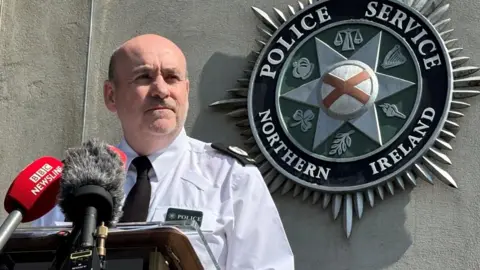 PA Media A man wearing a white PSNI shirt and black tie standing in front of microphones. There is a large PSNI logo on the wall behind him.