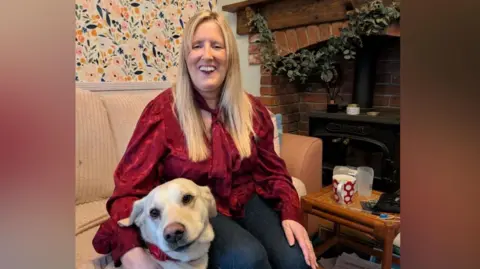 David Green The photo shows a woman with shoulder-length blonde hair smiling at the camera. She's wearing a red blouse and is sitting on a cream sofa. She's stroking a golden Labrador, which is sat to her left. 