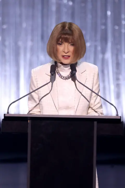 Getty Dame Anna Wintour in white dress and jacket, wearing a chunky necklace and sporting a sharp bob