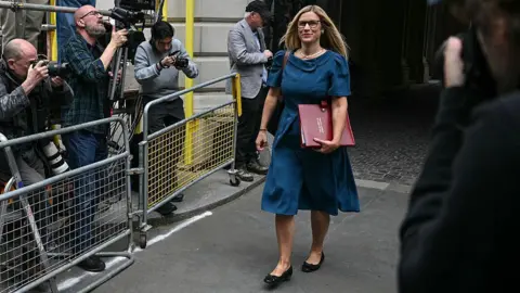 Getty Images Ellie Reeves carrying a red folder walks past photographers gathered behind barriers outside a government building.