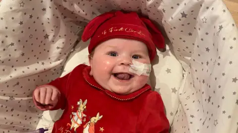 Supplied Six-month-old Amelia in a cot wearing a red baby grow and a matching red hat which says 'My First Christmas' in gold embroidery.