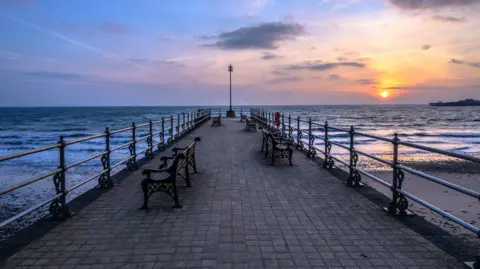 Richard Murgatroyd Looking directly down the pier, which has a circular end. There are benches on each side and a life float fastened to the railings. The paving is small square tiles. The sun is setting in the distance.