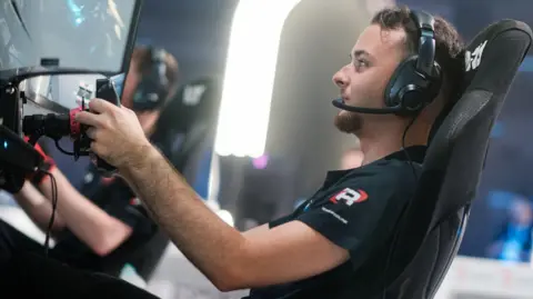 Esports World Cup Foundation Luke Bennett at the Esports World Cup in Saudi Arabia. Luke is a 19-year-old man with short brown hair and a short goatee beard. He wears a black T-shirt and headset and is pictured competing with a steering wheel, sitting in front of a screen