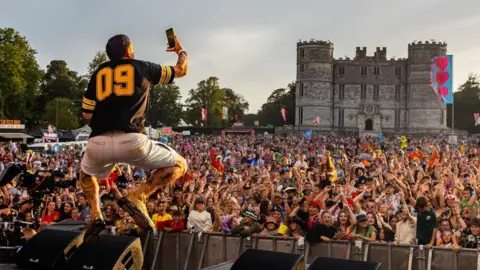 Camp Bestival Example, who has his back to the camera, is performing on stage. He is leaping into the air while holding his phone aloft in his right hand. Beyond him is a huge crowd of people, many of whom have their arms raised in the air. There is a large castle behind the crowd. 