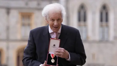 A 96-year-old man with wispy white hair holds a knighthood aloft to the camera. He is wearing a dark suit with a pink shirt and tie. 