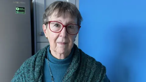 Christine Williams at their home. She is wearing a bluish-green top and cardigan and a pendant necklace. She has short hair and glasses and is looking at the camera with her hands behind her back.