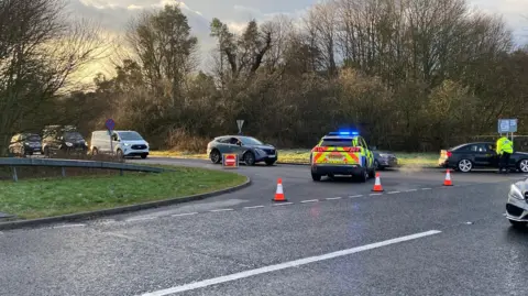 A police car is parked at the end of a road junction, and a police officer is stood diverting traffic at Denwick. There are cars in a line of traffic, with drivers waiting to speak with the police officer to be told where to go. There is a lot of greenery at either side of the road, and the sun is shining down on the cars from the left-hand side.