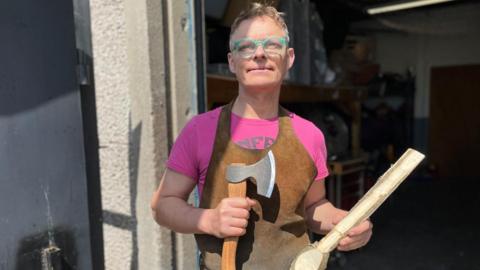 A man with green glasses and brown apron is holding an axe and a large wooden spoon