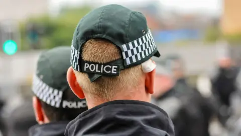 PSNI officers wearing baseball caps.  Two officers have been photographed from behind, with only the back of their heads and the collars of their dark jackets visible. 