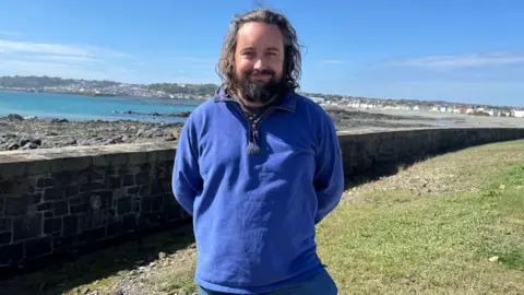 A man with long hear and a beard looking at the camera. He is wearing a blue three quarter zip