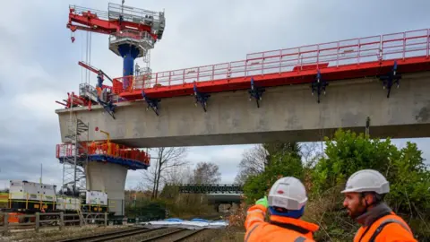 HS2 Ltd Two large viaducts being put into place over a railway line. Two people in orange hi-vis jackets are stood nearby.