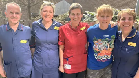 Royal United Hospitals Bath One man and five women in a mid-shot standing outside in sunshine and smiling to camera. Three are wearing blue medical uniforms, one is in a red uniform and the fifth is in a blue t-shirt with a cartoon picture on. There is a fence and greenery behind them