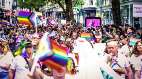 Members of the public with Pride flags walking along New Street in Birmingham during the Pride parade in 2024. The colours of the rainbow adorn each flag, lanyard and sign.