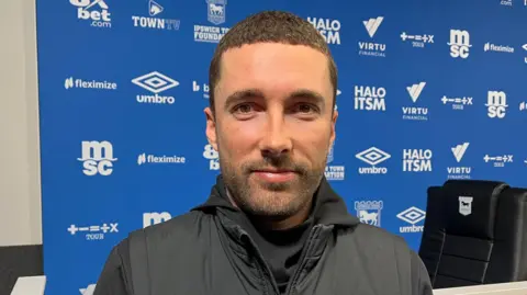 Tom Williams/BBC Joe Sheehan, the team's manager. He is looking straight at the camera. He has short black hair and a short black beard. He is wearing a black gilet and top. He is smiling. He is standing in front of a blue hoarding which is enblazoned with the names of sponsors.