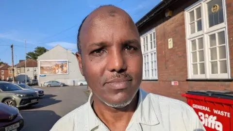 Nuh Sharif, a man with receding hair and a thin beard, is stood outside a social club in Barton Hill