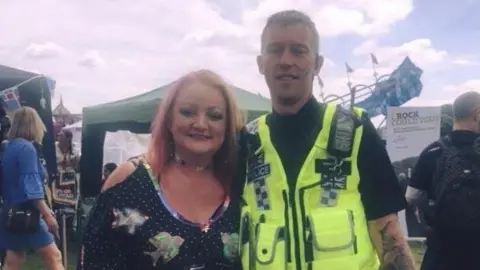 Gabby Rudge-Cox A couple smile at the camera at an outdoor event. The man is wearing a police vest, with the woman wearing a dress.