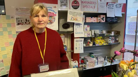 Amy Holmes/BBC A woman wearing a red sweater and a yellow lanyard stands behind a shop counter. Behind her are shelves filled with various products, notices, and promotional signs, including one labelled “Manager’s Specials.” Flowers are visible in the foreground to the right.