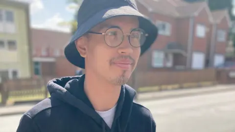 A man wearing a black hooded top and a black bucket hat with glasses, pictured with houses and a street behind him