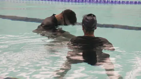 A man and woman are standing in a swimming pool. The man is putting his face into the water. He is wearing goggles