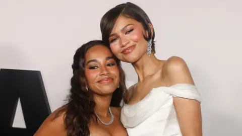 Getty Images Jordyn Curet and Zendaya pose, with their faces pressed against each other. Zendaya is wearing a white off-the-shoulder dress. 