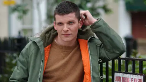Nigel Harman as Dennis Rickman in EastEnders in 2003. He has very short dark brown hair and wears a tan-coloured jumper with a green parka with orange lining and fur round the hood. He is resting his arm on a railing depicting the top of an Albert Square street sign.