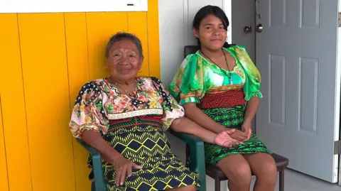 Magdelena and her granddaughter Bianka, in this weave, sat on plastic chairs in front of their new house. They are both wearing moolas with bright diamond -shaped design on them and looking at the camera directly. Their house has yellow and brown panels and a door behind them.