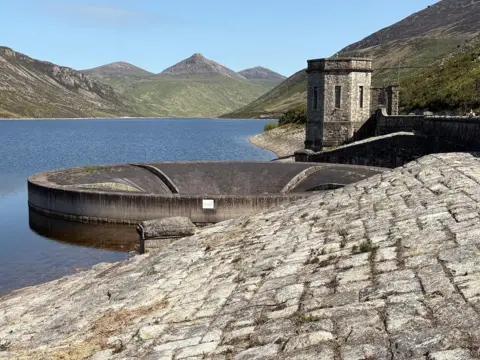 BBC Weather Watcher Pete’s Portal A body of water with a large concrete drain and tower in the middle. The mournes are pictured in the background, it is a clear sunny day.