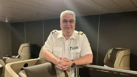 A man in a white shirt with and grey hair and glasses stands behind one of the sleeping pods which has its chair in the upright position
