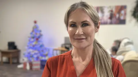 Care home manager Joeleen Logan poses for a photo in the residents' lounge. She has long, straight blonde haur and is wearing an orange top and a thin gold necklace. Reisdents are sitting in armchairs behind here 