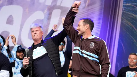PA Media Coventry City owner Doug King holding Frank Lampard's arm aloft, while holding a microphone on stage. Several members of the team are behind them, with a giant screen in the background
