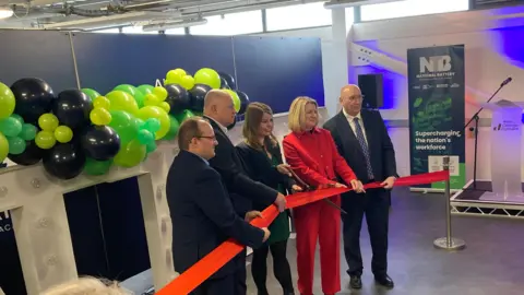 Five people holding a red ribbon as part of an opening ceremony. Left to right: a partially bald man in a dark suit, wearing spectacles; a bald man in a dark suit; a smiling woman with shoulder-length brown hair, wearing a dark green dress and black jacket, holding a pair of shears and about to cut the ribbon; a woman with blonde shoulder-length hair wearing a red suit, and a balding man wearing spectacles, a dark suit and a blue tie. Behind them is a strand of green and black balloons