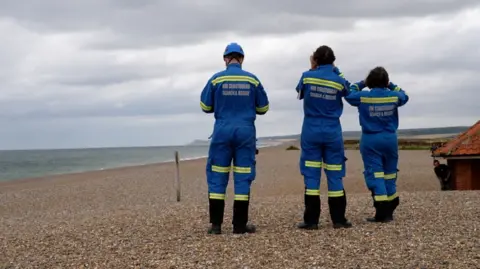 Three coastguard rescue officers during a search operation look out towards the sea on a shingle beach along the north Norfolk coast.