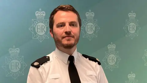 Sup Int Phil Mullally has short brown hair and a brown beard. He is wearing a white police shirt and a black tie.