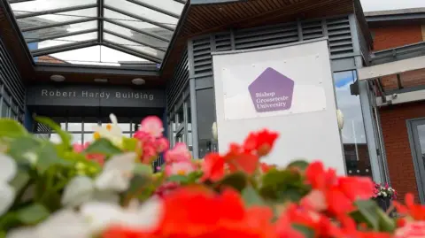 A purple pentagram sign for Bishop Grosseteste University is on the front of a building, in front of that is a selection of summertime flowers in bloom. From reds, pinks and whites, the flowers appeared blurred against the focused sign in the background.