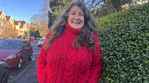 Kitty Wilson smiling, she has long, curly dark hair and is wearing a red knitted jumper. She is pictured outside, standing on the pavement next to a hedge and a row of parked cars.