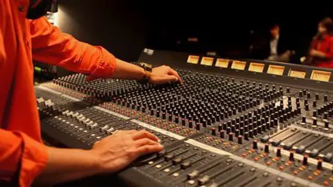 BBC Man in orange shirt twisting black knobs on a grey recording desk