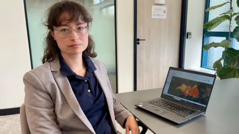 A woman in a smart grey blazer and navy blouse sat at a desk. There is a laptop on the desk showing a satellite image of a wildfire. There are windows and plants in the background.
