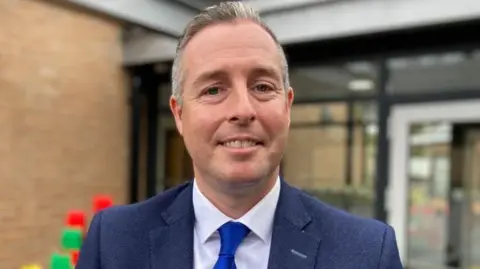 BBC Paul Givan is wearing a bright blue tie and a navy blazer. He has short brown and grey hair. 
