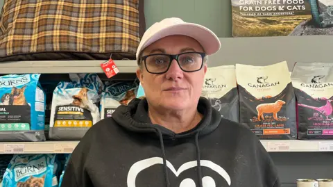 Pet shop owner Sarah Le Roux, a woman in a pink hat, glasses and black hoody is looking at the camera. Behind her on shelves are bags of pet feed.