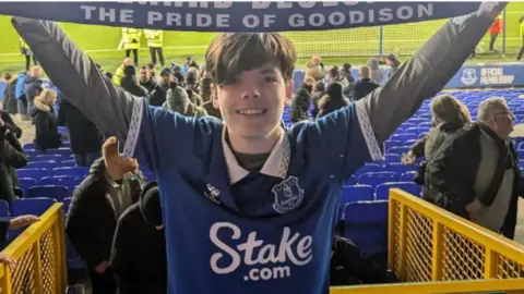 Handout A smiling Mackenzie holds an Everton scarf over his head with the Goodison Park pitch behind him