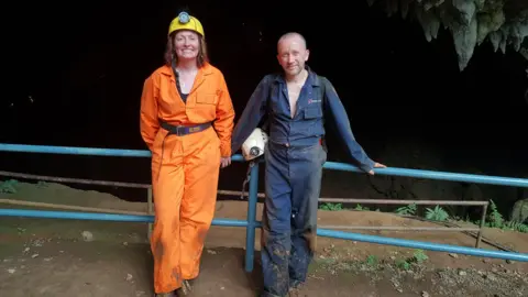 Mike Clayton in blue overalls holding a white helmet is pictured with his wife Emma in an orange overall jumpsuit. They are standing at the entrance to Tham Luang 