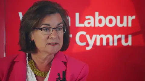 PA Media Eluned Morgan is to the left of the image, wearing a pink suit with a white shirt and gold necklace. She has short dark hair in a bob and glasses on. Behind her is a sign that says Labour and Cymru underneath is white font.
