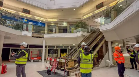 Stack The inside of a former shopping centre, with a number of people standing in it, wearing hi-vis jackets and hard hats. There is an escalator and shop fronts. There is a balcony. The building is white.