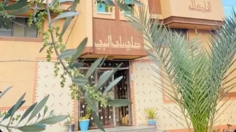 Ashraf Abo Taha An image of a house painted in different shades of beige, with distinctive white and beige tiles around the entrance and plants in front