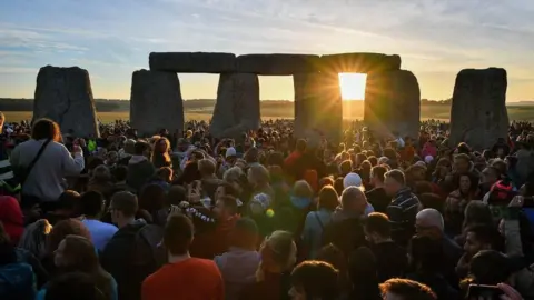 PA Media A large crowd of people gathered around Stonehenge as the sun rises over the horizon. The sunlight is bursting through one of the stone archways.