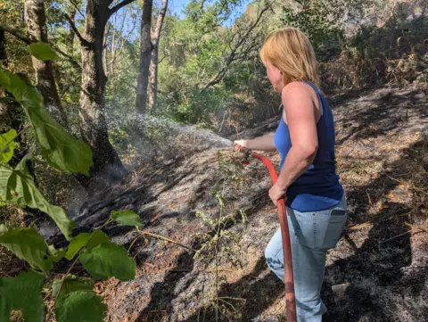 Tommy Udall Donna Oldbury-Thomas pictured holding a hose that has water coming out of it. The water is going onto the ground. The ground is scorched black and there are trees ahead of her.