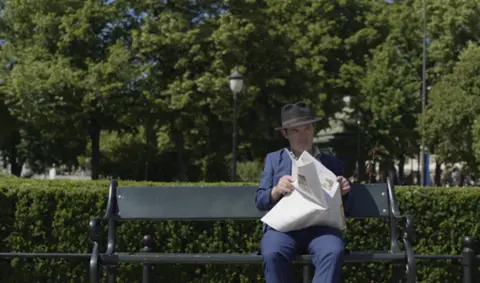 Svein Andre Hofsø sitter på en parkbenk. Han har på seg en marineblå dress og holder en delvis åpen avis på fanget. Han har en hatt på hodet og ser ut til å se til venstre. Det er trær og busker bak ham